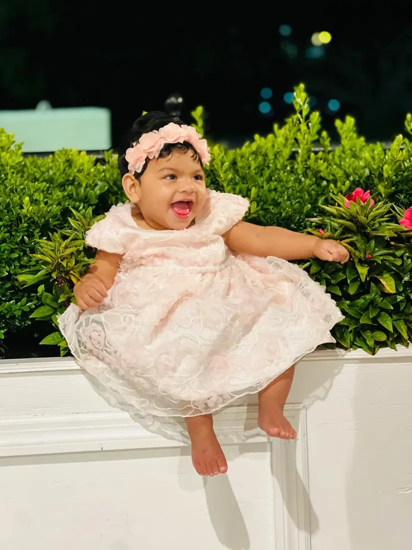 A smiling child in a pale pink dress surrounded by greenery and flowers