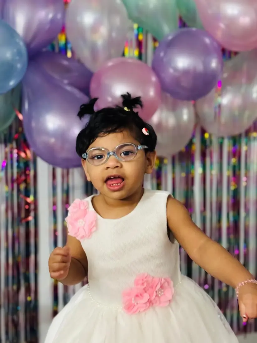 A child in a white dress with pink flowers in front of pastel balloons