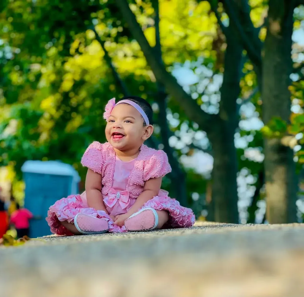 A smiling child in a pink dress sitting outdoors beneath green trees